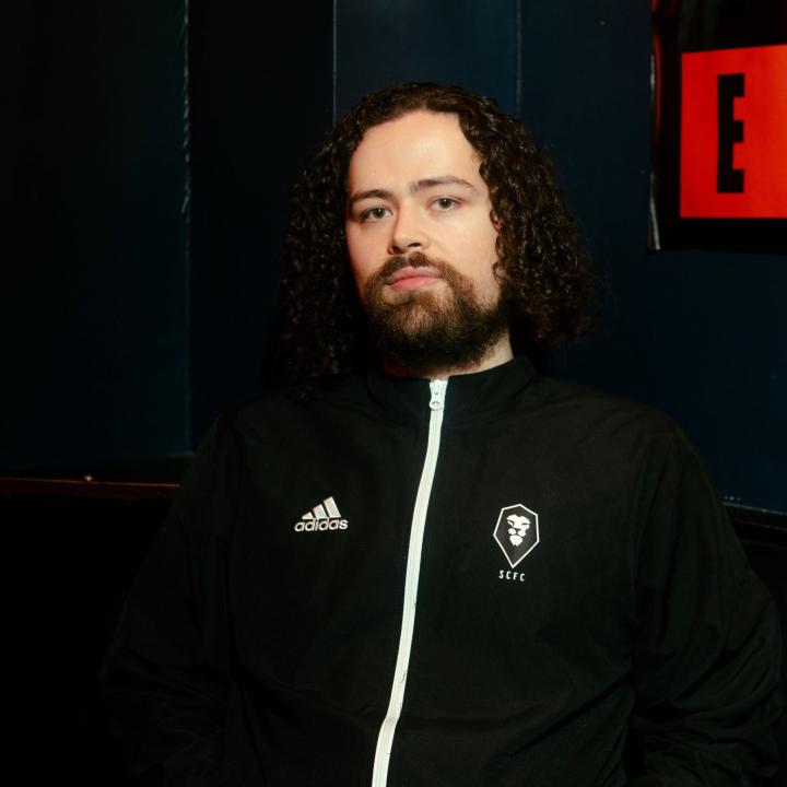 Portrait of a person with long, dark curly hair and a full beard, looking directly at the camera with a neutral expression. They are wearing a black Adidas Salford City FC tracksuit jacket with a white zip and club crest, against a dark blue background.
